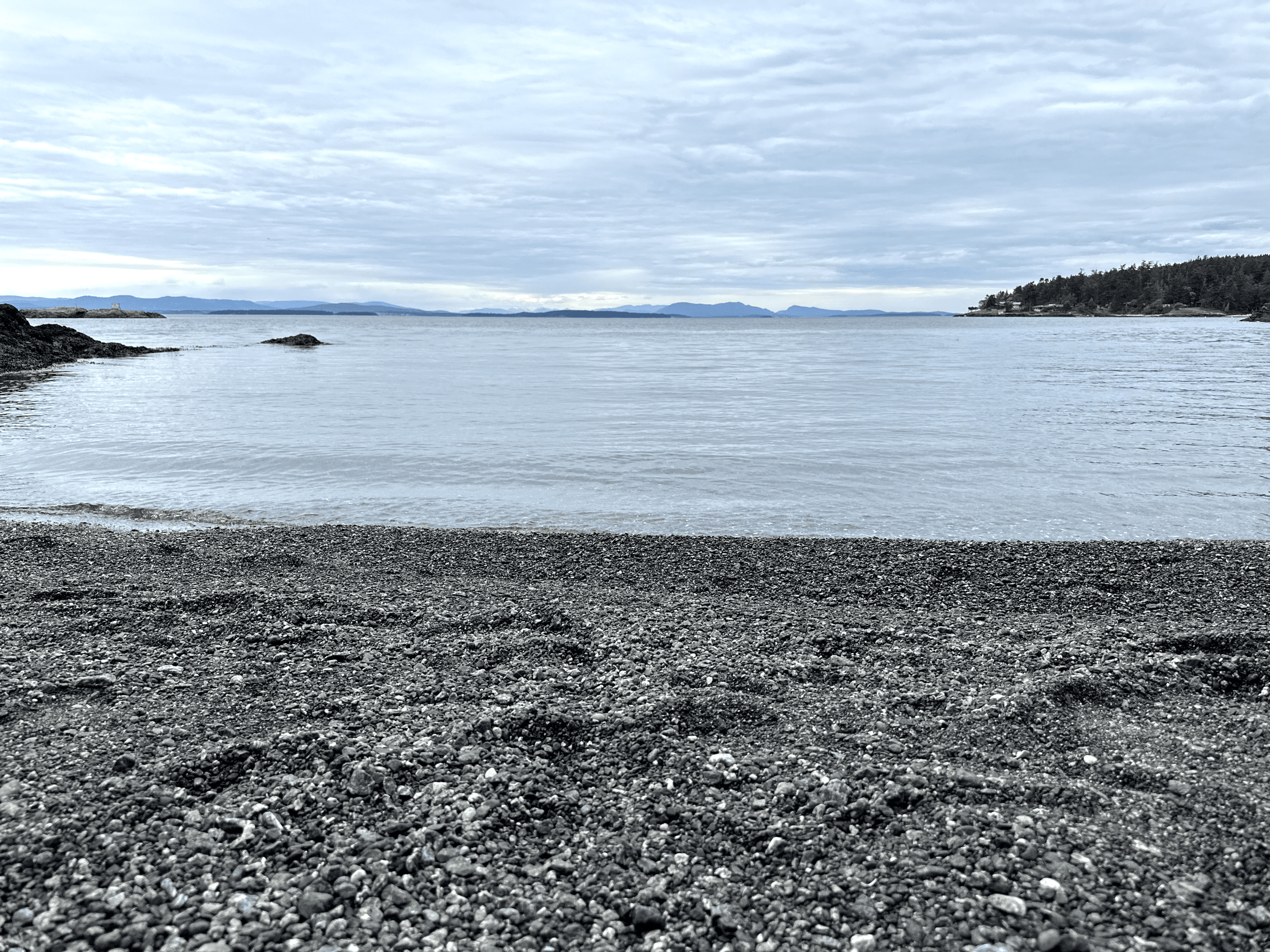 Tranquil beach scene with pebbles, calm water, distant mountains, and scattered clouds.