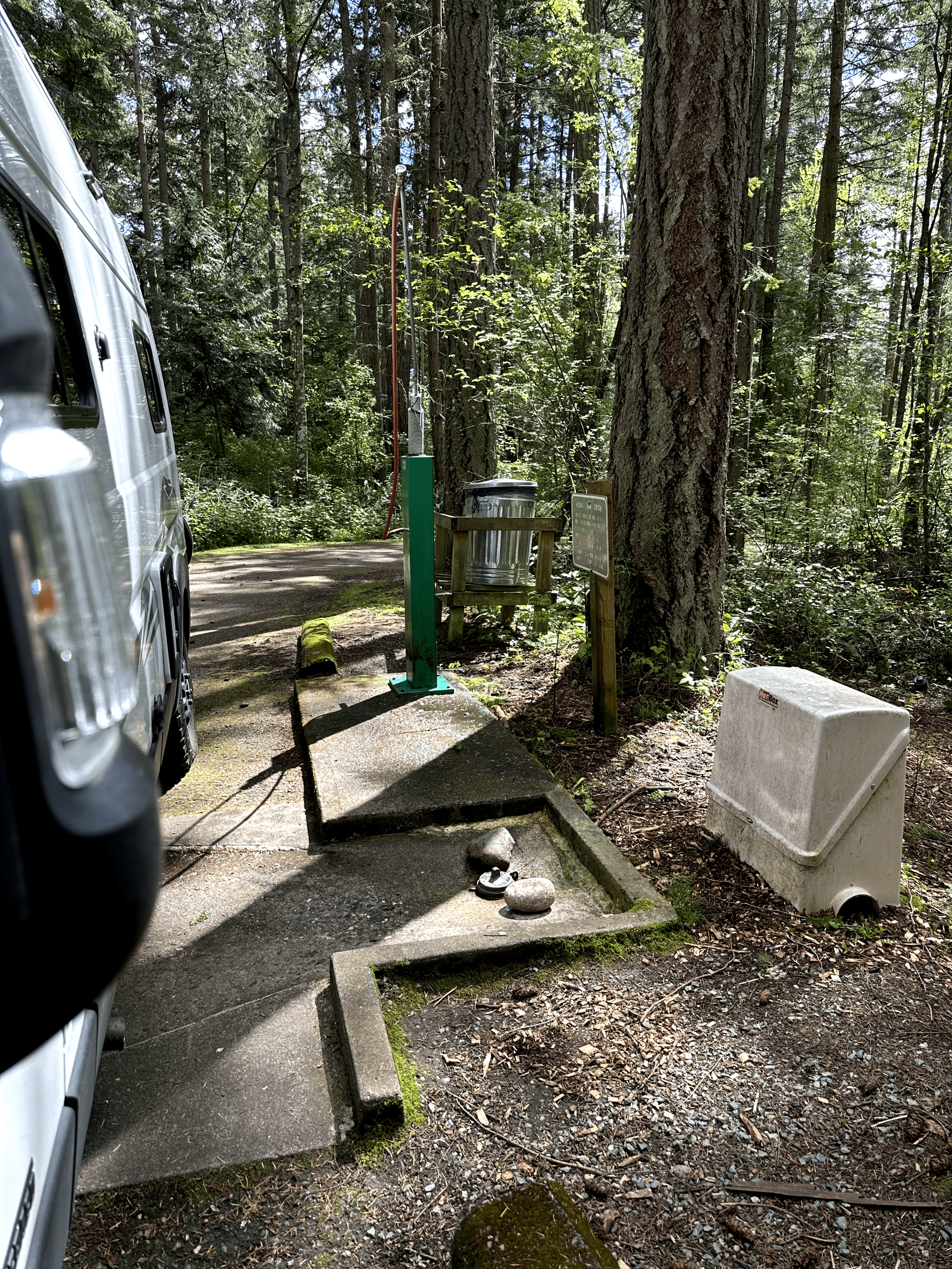 Campsite in forest with van, recycling bin, and signboard for campers.