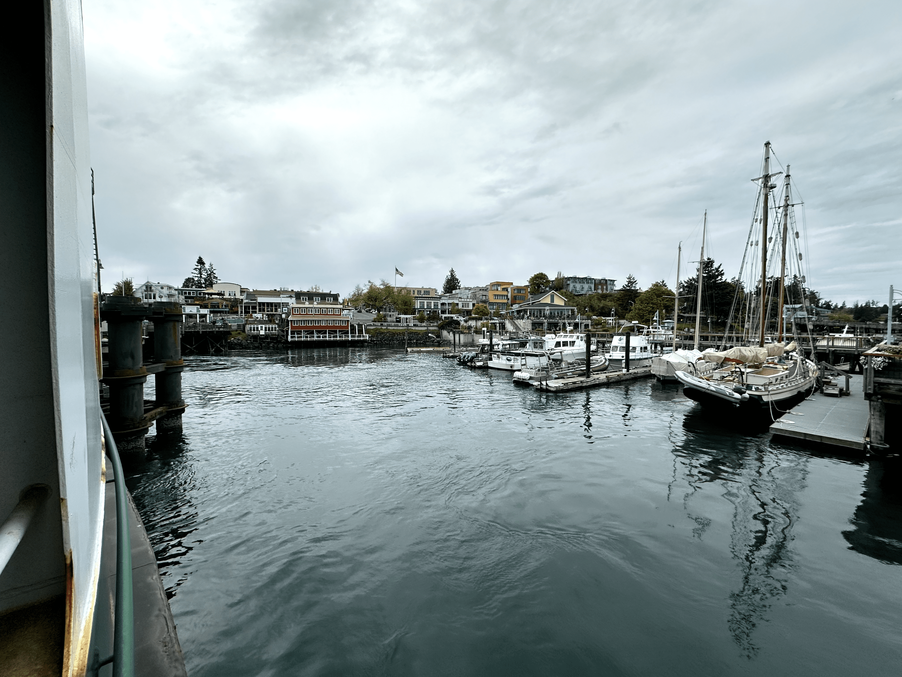Tranquil harbor with boats moored, sailing ship, overcast sky, waterfront buildings, and greenery.